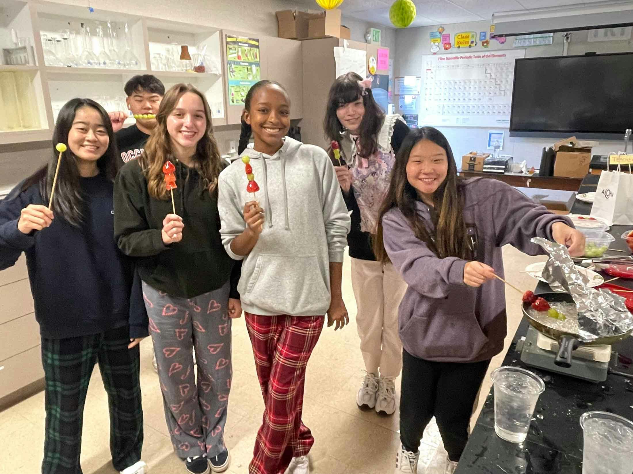 Six teens in a kitchen holding skewers of fruit, some coated in a candy glaze