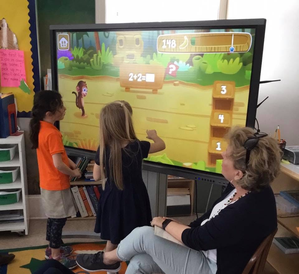 Two children interacting with a large screen in a classroom, person watching