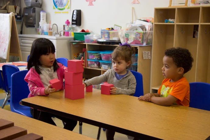 Three children playing with red building blocks at a table in a classroom
