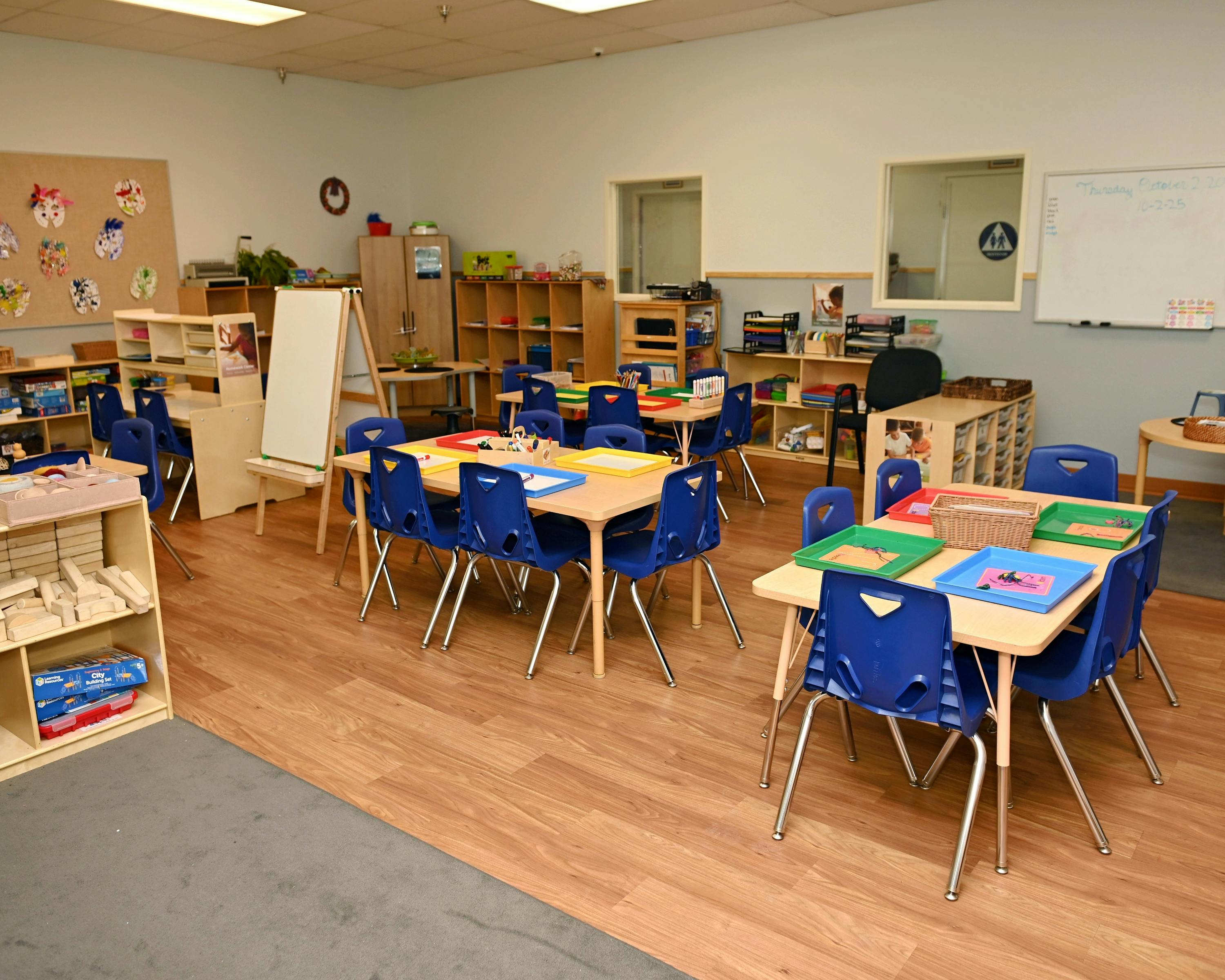 Classroom with small tables and blue chairs, shelves, and a whiteboard