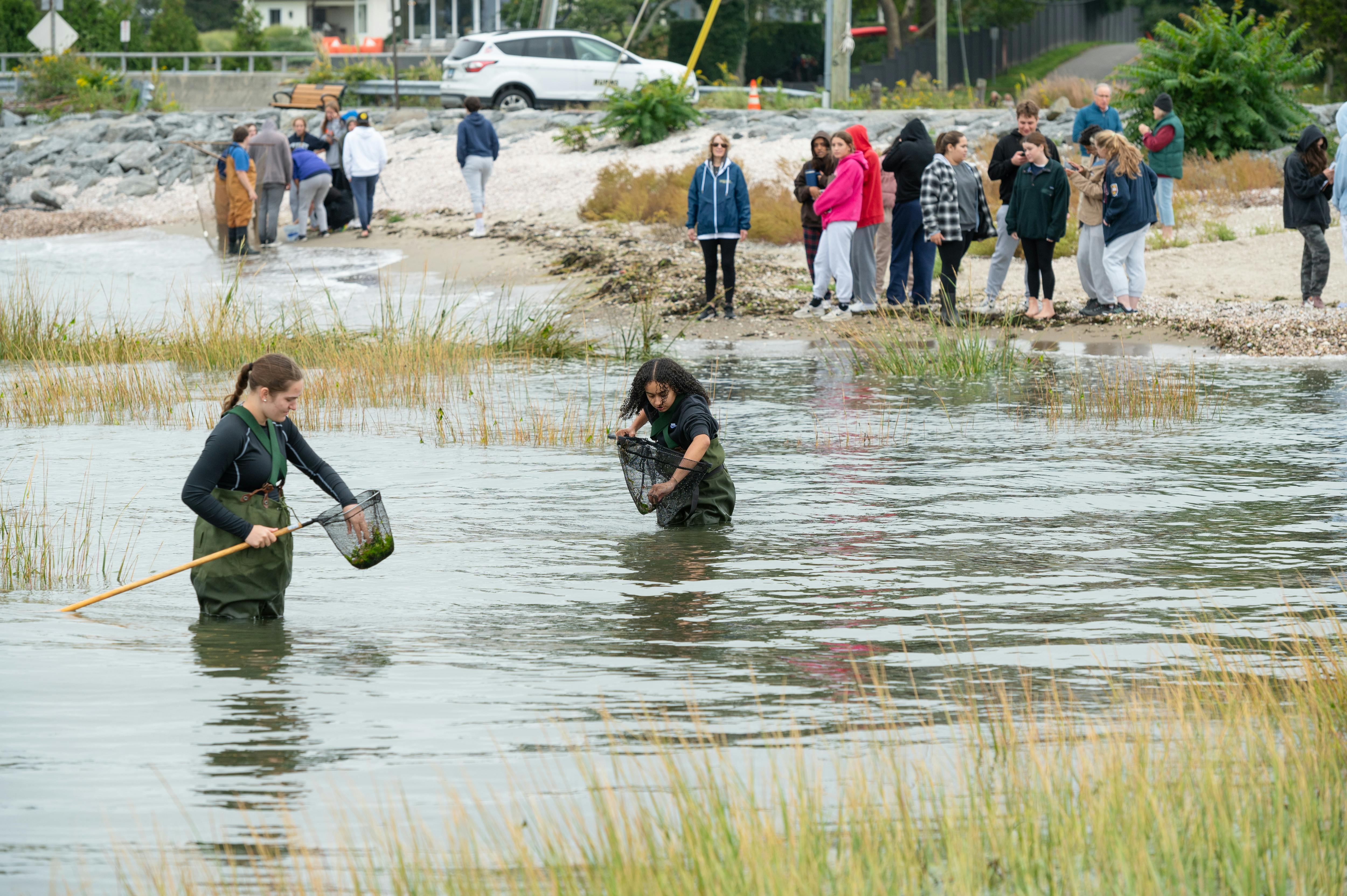 Two people in waders in water, one holding a net, with people on shore