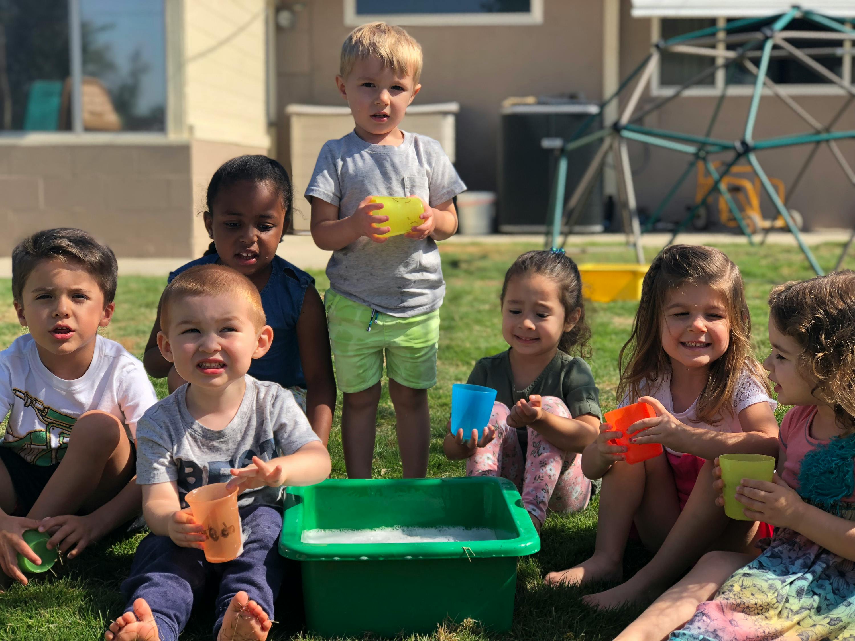 Group of children sitting on grass with cups and a tub of water