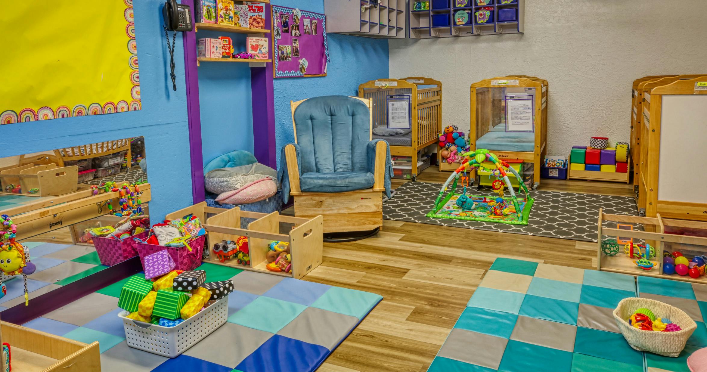 A childcare room with cribs, play mats, shelving with toys, and a rocking chair