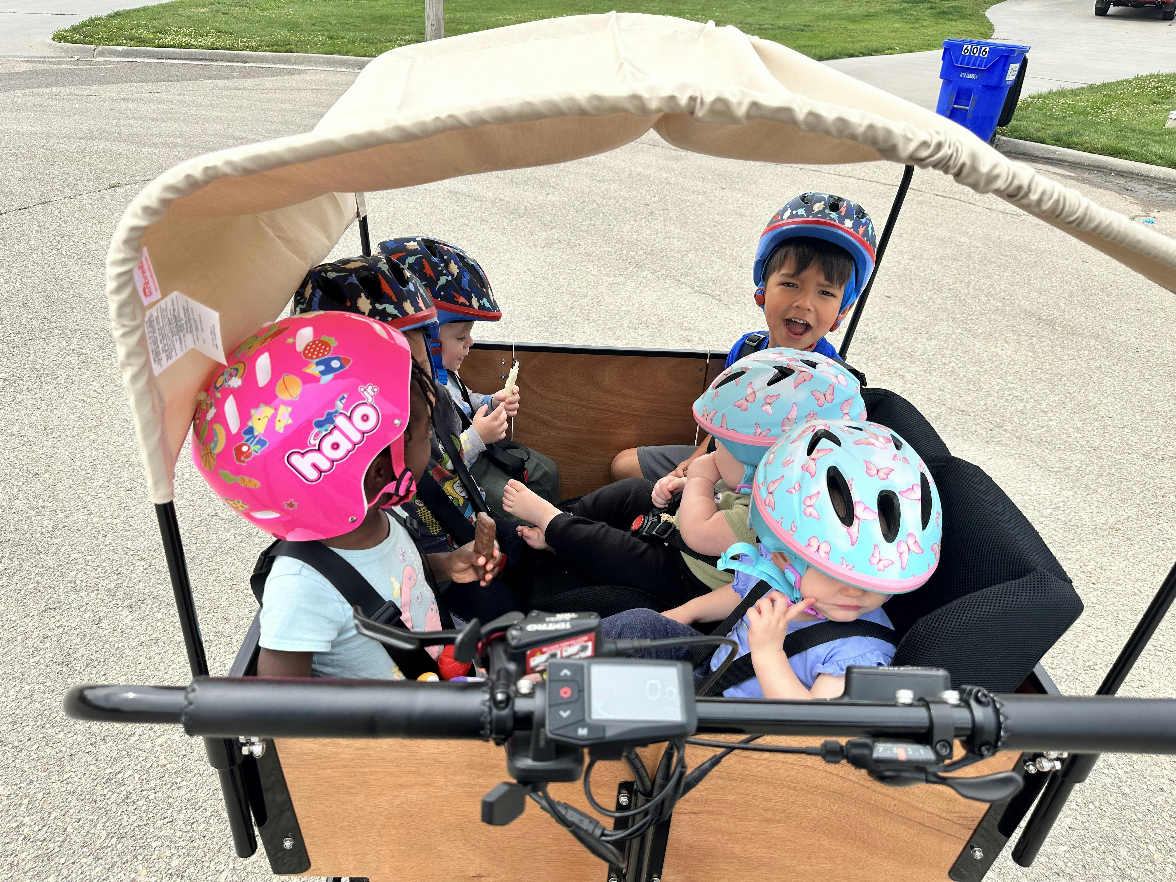 Five children in helmets seated under a canopy in a cargo bike