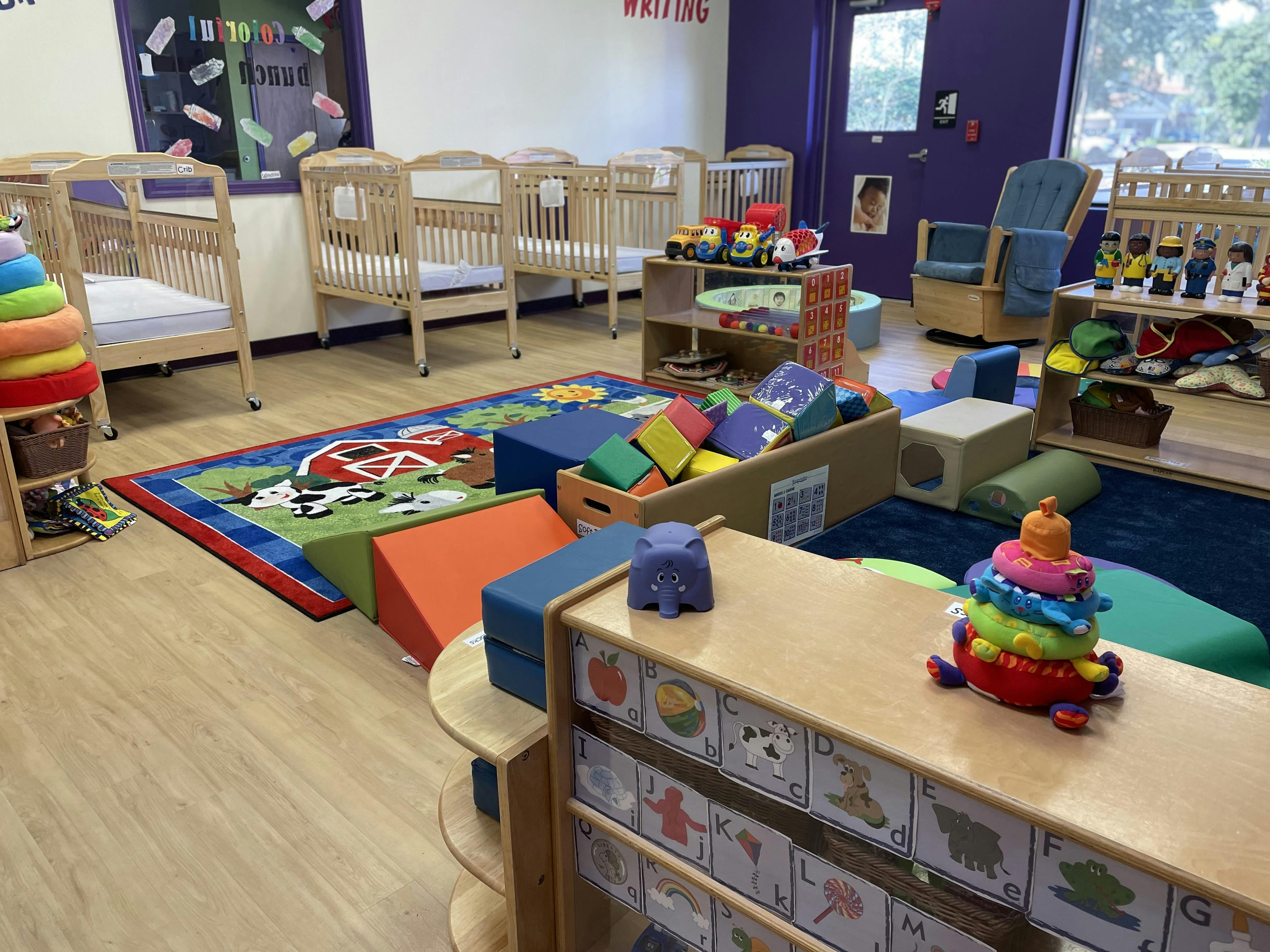 Classroom with multiple cribs, a play rug, colorful soft blocks, and toys
