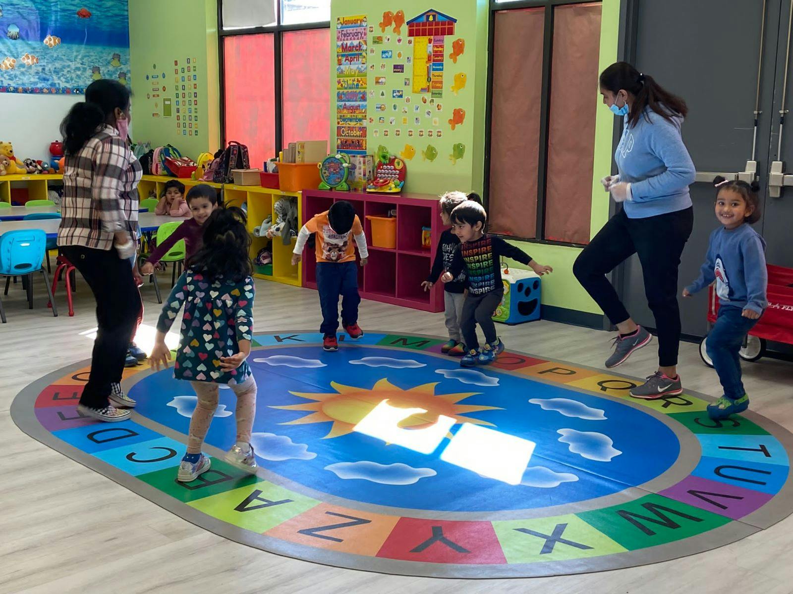 Two adults and multiple children playing on a colorful alphabet rug in a classroom