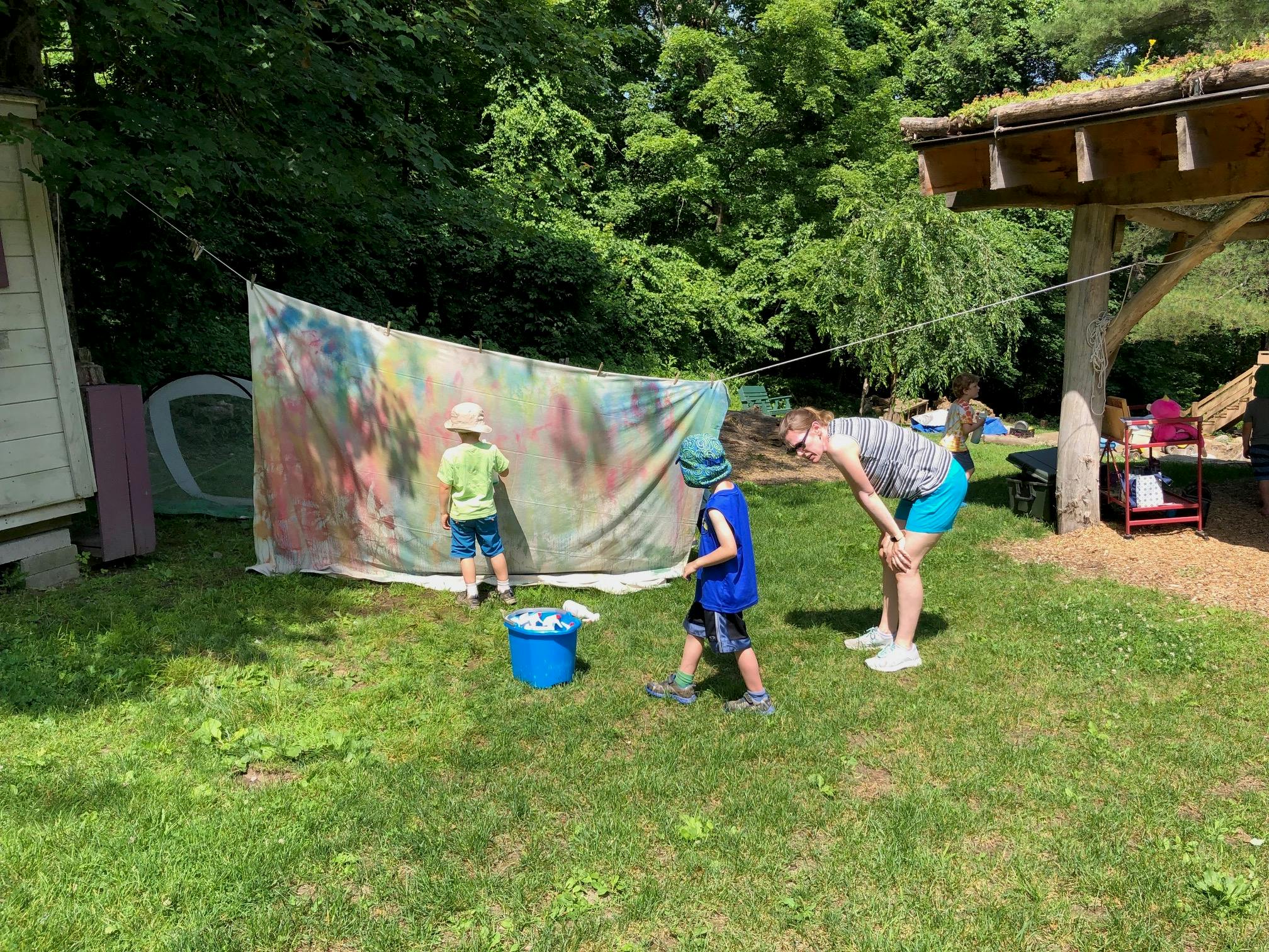 Two children painting on a large fabric sheet outdoors with an adult nearby