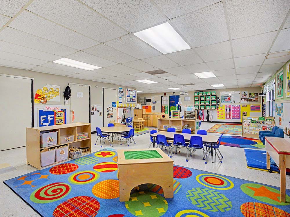 Classroom with small tables and chairs, colorful rugs, and shelves with toys