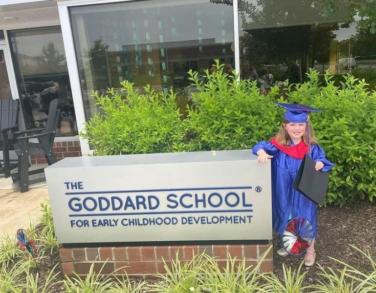 Child in graduation cap and gown standing next to "The Goddard School" sign