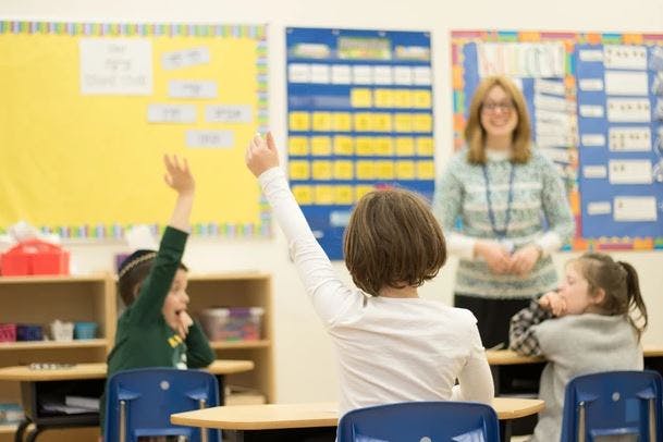 Children raising hands in a classroom with an adult standing in the background