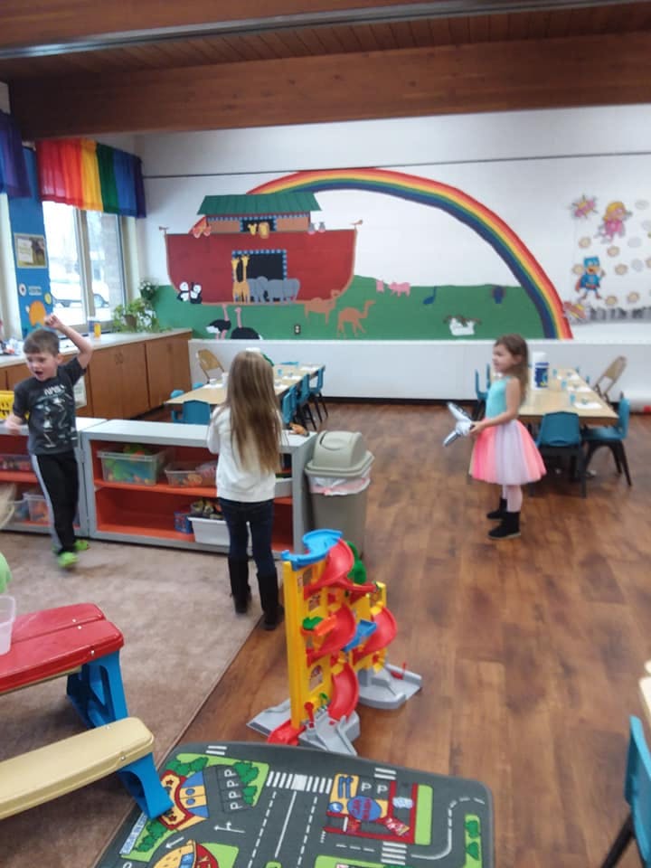 Three children in a classroom with a mural of an ark and rainbow
