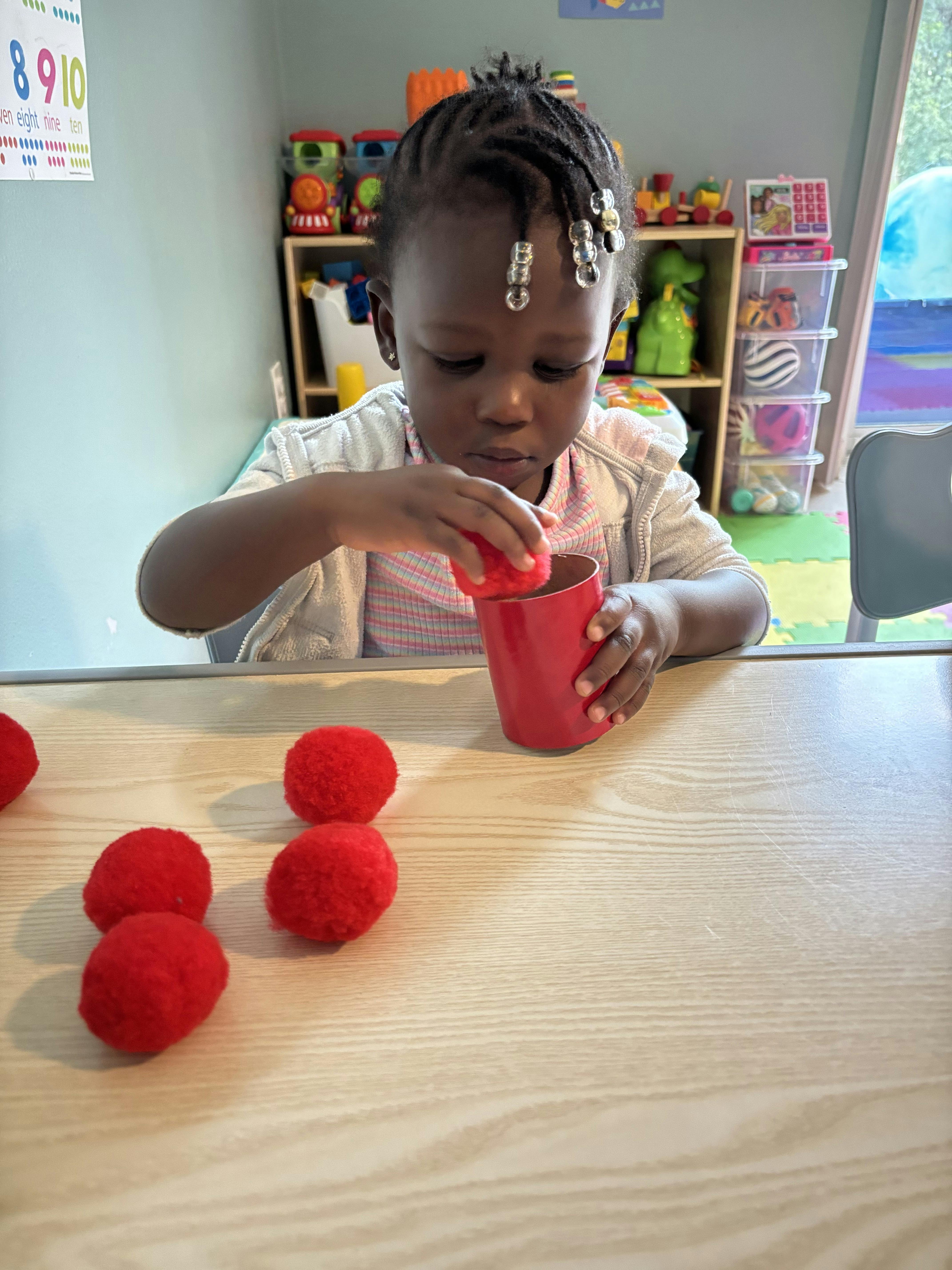 Child putting a red pom-pom into a red cup on a table