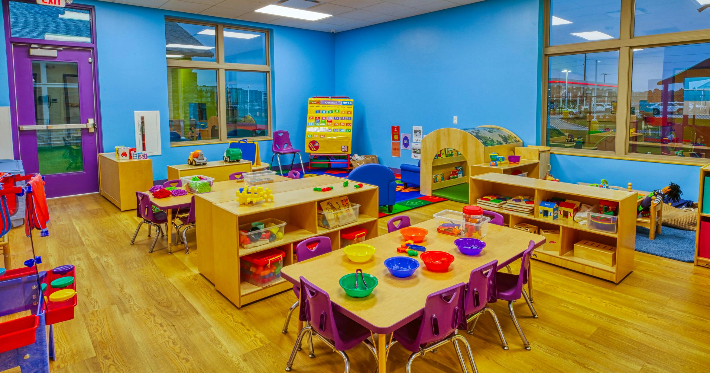 Colorful classroom with small tables and chairs, shelves with toys, and an October 2025 calendar