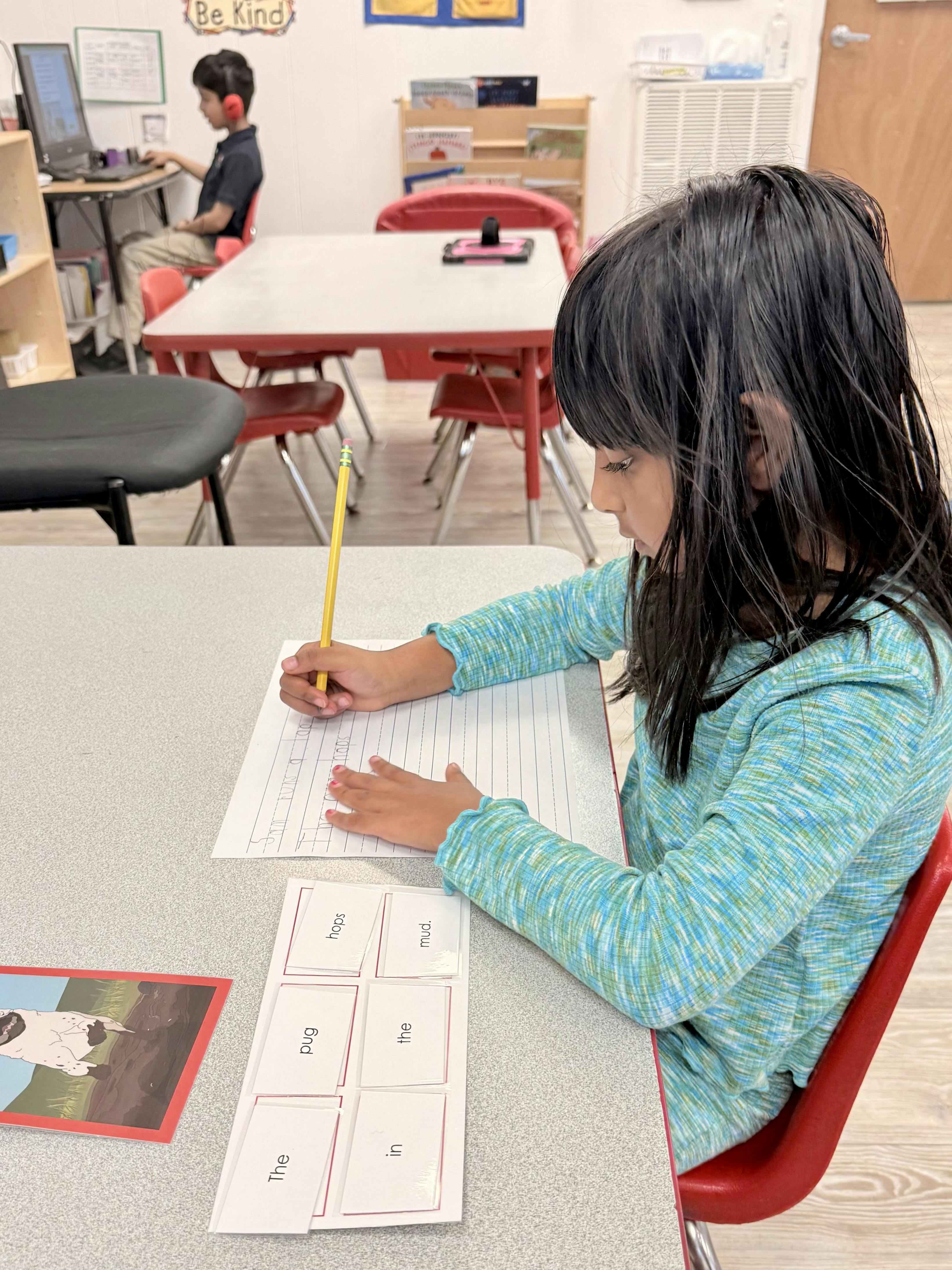 Child writing on a lined paper with a pencil at a desk in a classroom