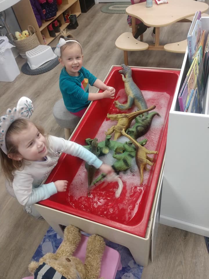Two children playing with dinosaur toys in a sensory bin with water and bubbles