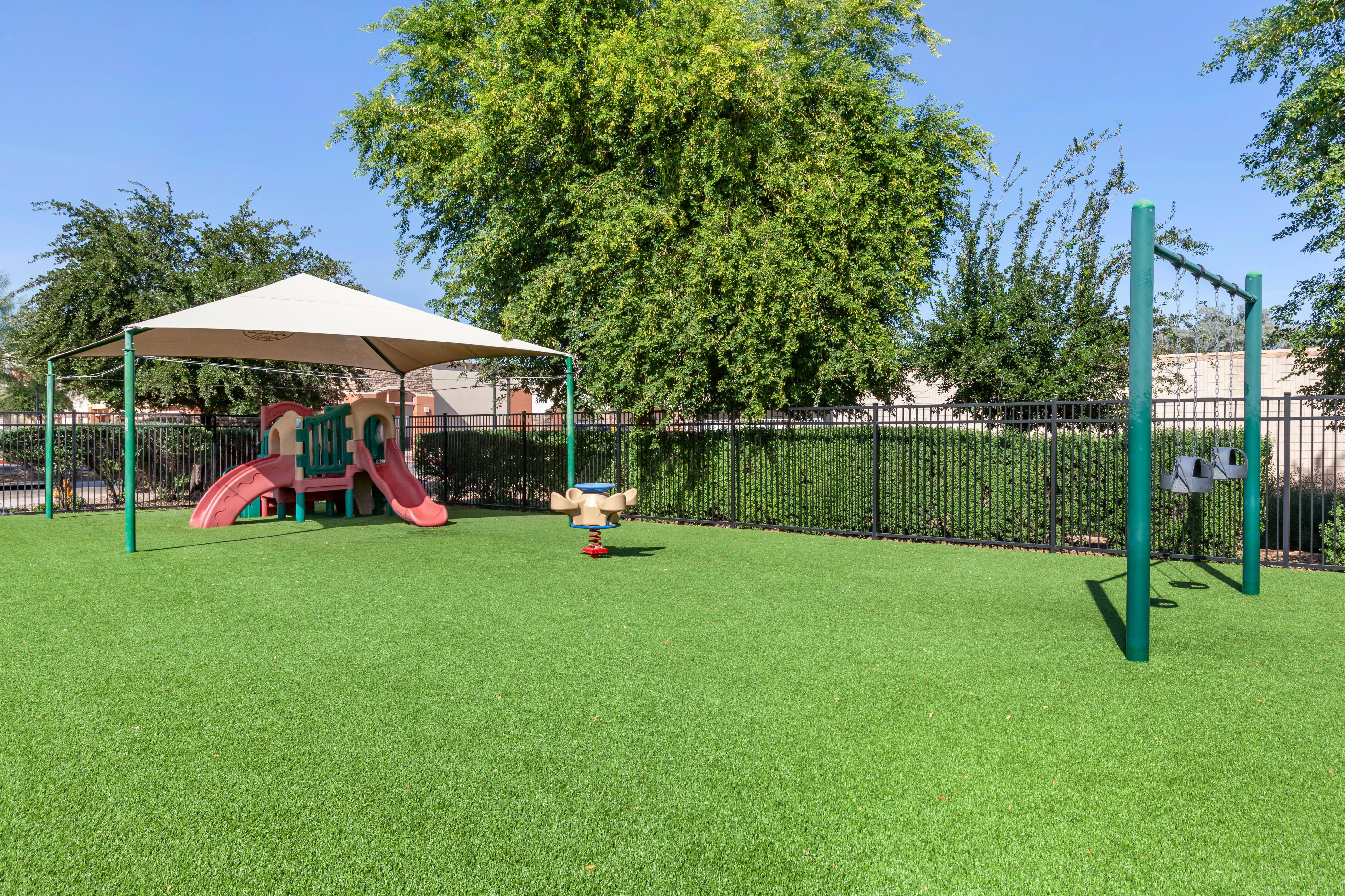 A playground with artificial grass, slides, a spring rider, and swings under a shade