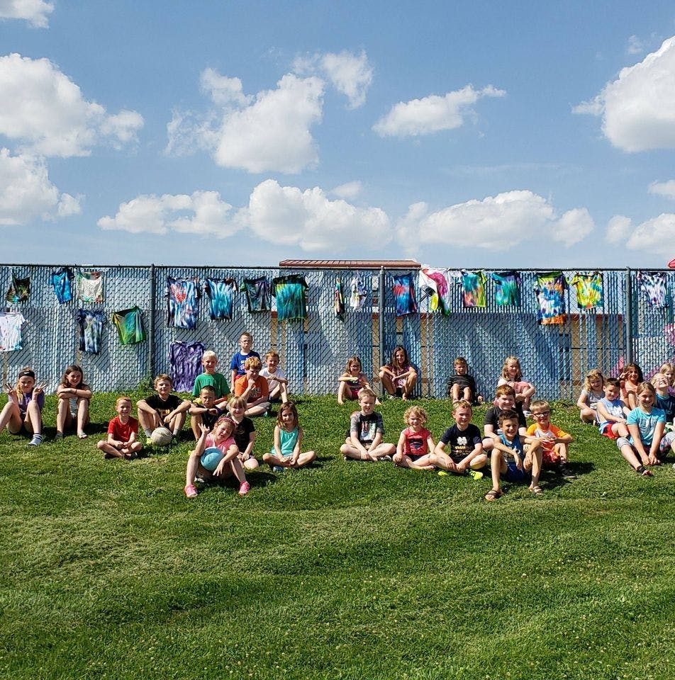 Group of children sitting on grass in front of a fence with hanging shirts